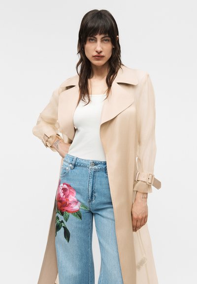 Femme aux cheveux foncés portant un trench-coat beige, un haut blanc et un jean bleu avec un grand imprimé de fleur rose sur la cuisse gauche.