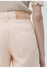 Denim hlače v breskevni barvi z zadnjim žepom in kožnim označevalnikom v svetlo rjavi barvi z napisom "MNG DENIM." Strukturirana pletena majica se nosi na pasu.