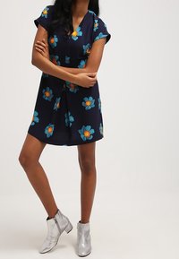 Mujer vestida con un vestido azul marino con grandes flores azules y botas plateadas hasta el tobillo, de pie con los brazos cruzados contra un fondo sencillo.