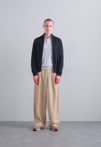Juodas zippered švarkas uždėtas ant šviesiai pilkos polo marškinėlių, derinamas su laisvomis smėlio spalvos kelnėmis ir rudos spalvos sportiniais bateliais. Paprastas dizainas su neutraliosiomis spalvomis.