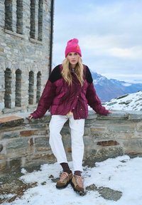 Bordový vypchávkovaný kabát s čiernymi plecami, biele nohavice, hnedé turistické topánky s ružovými šnúrkami a jasne ružová čiapka. Snehový vonkajší prostredie.