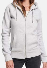 Sudadera con capucha gris claro con cremallera, hecha de tela suave, que cuenta con capucha, cordones y dos bolsillos delanteros. Puños y dobladillo acanalados para un ajuste ceñido.