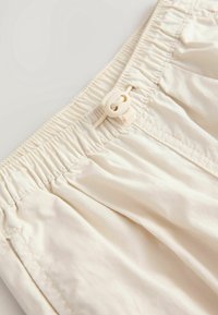 Gros plan de la ceinture d'un pantalon couleur crème avec bande élastique et cordon réglable avec verrou.