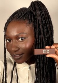 Brun leppestift i rektangulær emballasje, merket "575 THE LIPSTICK," holdt av en person med lange svarte flettehår, iført en blank beigefarget jakke.