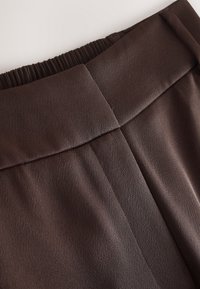 Plan rapproché d'un pantalon en tissu marron foncé présentant une large ceinture avec un fronçage élastique à l'arrière et un détail de couture centrale.