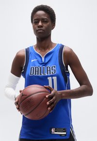 Γυναίκα παίκτρια μπάσκετ που φοράει μπλε φανέλα των Ντάλας, κρατώντας μπάλα μπάσκετ με τα δύο χέρια μπροστά σε απλό φόντο