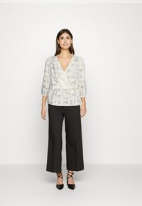Blusa branca envolvente com padrões abstratos cinzentos, mangas de três quartos, cintura ajustada, combinada com calças largas pretas e sapatos pretos de bico apontado.