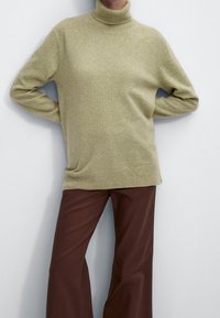 Hellgrüner Rollkragenpullover aus weichem Stoff mit lockerem Schnitt, kombiniert mit braunen Hosen mit weitem Bein. Glatte Textur, minimalistische Details.