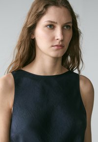 Junge Frau mit langen, welligen Haaren, die ein ärmelloses schwarzes Oberteil trägt und mit neutralem Ausdruck leicht seitlich blickt, vor einem schlichten Hintergrund.