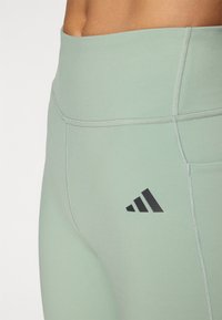 Világoszöld sportleggings, széles derékpánttal, lapos varratokkal és fekete Adidas logóval a bal combján.