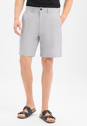 Man draagt lichtgrijze gestreepte korte broek en zwarte sandalen, staat met één hand in de zak tegen een effen achtergrond.