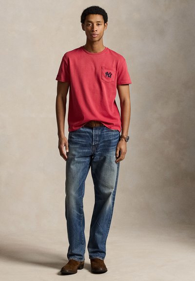 Homme portant une chemise rouge à manches courtes avec un logo sur la poche, un jean bleu, une ceinture marron et des bottes marron, debout devant un fond neutre.