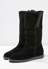 Stivali alti fino al ginocchio in camoscio nero con pannelli elastici sui lati, suola piatta in gomma e cuciture visibili lungo le giunture. Fodera morbida.