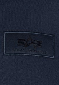 Material de culoare albastru închis, cu o pată texturată neagră, ce prezintă textul în relief "ALPHA INDUSTRIES" și un logo stilizat, de formă rectangulară.