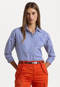 Blauw-wit gestreepte overhem shirt met kraag, met lange mouwen opgerold. Het shirt heeft een geborduurd detail op de linkerkant van de borst.