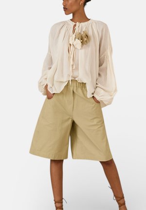 Donna che indossa una blusa crema larga con nappine floreali e pantaloncini beige larghi fino al ginocchio, con le mani nelle tasche, su uno sfondo semplice.