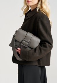 Borsa a tracolla in pelle grigia con un design strutturato, dotata di una fibbia prominente, una tracolla removibile e una finitura testurizzata, tenuta sotto il braccio.