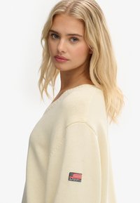 Blondynka o niebieskich oczach, ubrana w kremowy sweter z dzianiny z małą łatką logo Superdry w kolorze czarnym, czerwonym i białym na lewym rękawie.