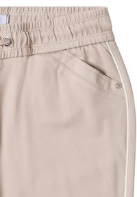 Béžové sportovní kalhoty s elastickým pasem, boční kapsou a bílým lemováním podél švů. Hladká textura látky.