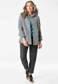 Grau meliertes Wollmischjackett mit Knopfverschluss, kombiniert mit einem blauen Strickpullover, dunkelgrauen Hosen und metallischen Sneakers mit schwarzen Schnürsenkeln.