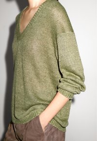 Olijfgroen gebreid sweater met een V-hals, ontspannen pasvorm en opgerolde mouwen. De textuur is los en ademend. Gecombineerd met een paarse broek.