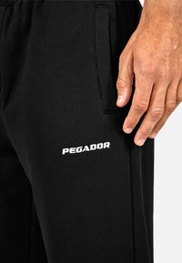 Zwarte joggers van zacht materiaal met een losse pasvorm, voorzien van zijzakken en een wit "PEGADOR" logo op de dij.