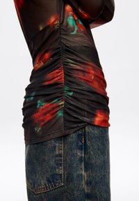 Czarna, przejrzysta bluzka z marszczonym wzorem w kolorowy, abstrakcyjny motyw w odcieniach czerwieni, zieleni i żółci; zestawiona z niebieskimi dżinsami.