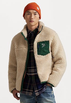 Uomo con berretto di lana arancione, giacca in sherpa crema con tasca verde con zip, camicia a quadri e jeans blu strappati, in piedi con una mano in tasca.