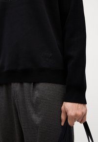 Felpa nera con una texture liscia, con scollo rotondo e un discreto dettaglio del logo. Pantaloni a quadri grigi completano l'outfit.