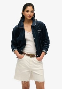 Veste bomber en velours marine avec accents blancs, fermeture éclair à l'avant, manches à côtes et détail brodé. Portée par-dessus un haut avec dentelle blanche et un short blanc.