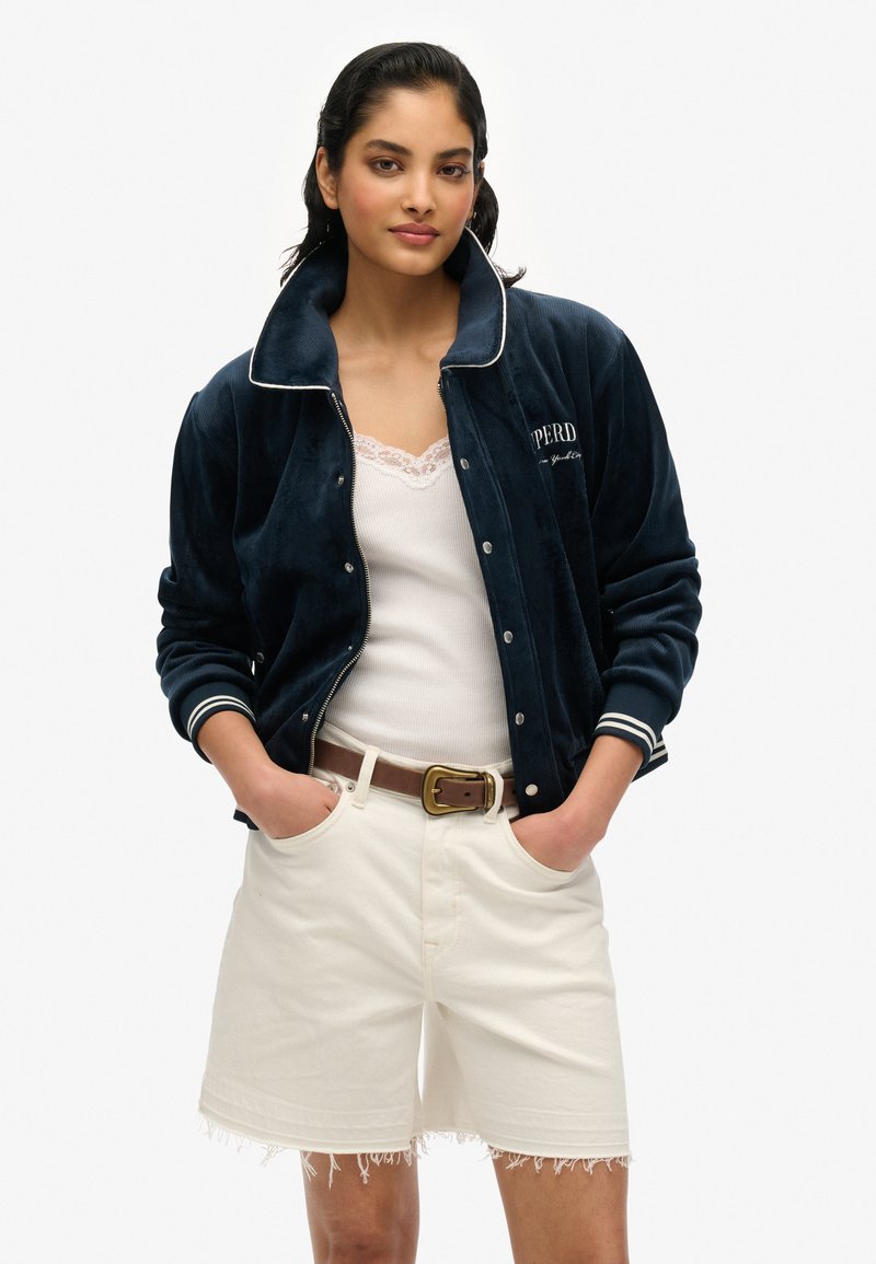 Veste bomber en velours marine avec accents blancs, fermeture éclair à l'avant, manches à côtes et détail brodé. Portée par-dessus un haut avec dentelle blanche et un short blanc.