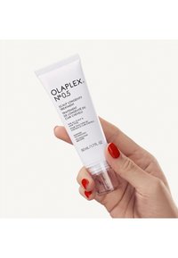 Olaplex No. 0.5 hoofdhuidbehandeling, in een witte tube met een doorzichtig onderkant. Zwarte tekst, 50 ml formaat. Hand met rode nagellak die de tube vasthoudt.