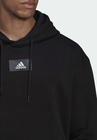 Uomo che indossa un hoodie nero Adidas con cordini, caratterizzato da un logo Adidas rettangolare sul petto, mostrato dalla spalla alla bocca.