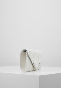 Pochette blanche avec un rabat en dentelle texturée et un corps en cuir lisse. Le design comprend un sommet triangulaire et des bords arrondis pour une forme élégante.