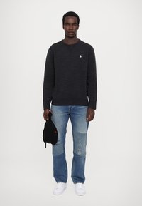Mørk grå hettegenser med ribbestrikket hals og mansjetter, brukt med lyseblå slitte jeans og hvite sneakers, holder en svart caps.