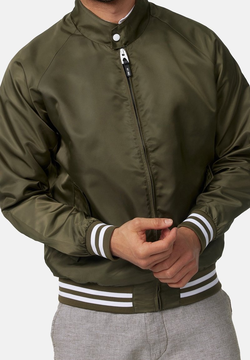 Homme portant une veste bomber vert olive avec poignets et ceinture côtelés rayés blancs, mains croisées devant, assortie à un pantalon gris.