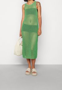 Abito maxi verde lavorato a maglia con scollatura a barca e spacchi laterali, abbinato a una borsa color crema e sandali platform beige.
