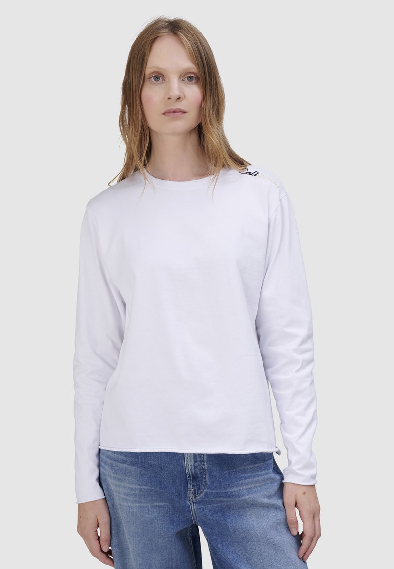 Weißes langärmliges Hemd mit rundem Ausschnitt, minimalistischem Design und einem dezenten bestickten Detail an der Schulter. Kombiniert mit blauen Jeans.