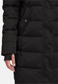 Sort polstret frakke med en quiltet tekstur, der har en sidelomme og et design med manchetter på ærmerne. Materialet virker mat og glat.