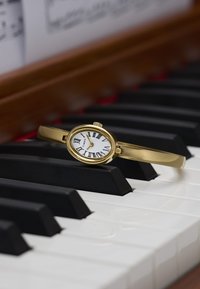 Gouden ovalen Timex-horloge met witte wijzerplaat, Romeinse cijfers en een gladde band die rust op zwart-witte pianotoetsen.