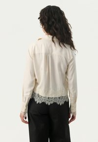Cremefarvet bluse med blondekant i nederste kant og ved ærmerne, glat tekstur, knaplukning foran og en afslappet pasform, stylet med mørke bukser.