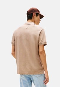 Polo de manga corta beige con tela texturizada y cuello clásico, combinado con vaqueros azul claro y una gorra marrón.