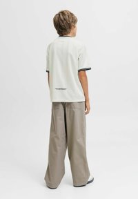 Niño con cabello castaño claro de pie de espaldas, vistiendo una camisa blanca de manga corta con ribete negro y pantalones anchos beiges, frente a un fondo sencillo.