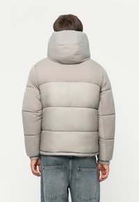 Lys grå dunjakke med høy krage, med quiltet design og elastiske mansjetter, brukt over blå jeansbukser.