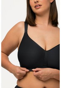 Sostén inalámbrico negro con tejido suave, tirantes ajustables y diseño de escote en V. La persona que lo lleva puesto está ajustando la banda debajo del busto.