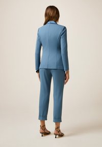 Completo blu su misura con blazer aderente e pantaloni cropped, realizzato in tessuto morbido. I dettagli notevoli includono maniche arrotolate e tacchi con cinghiette.