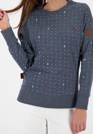 Pull gris avec des points blancs et de petites touches de rouge, motifs d'ancre de bateau, bords côtelés aux poignets et à l'ourlet, avec un patch en cuir marron.