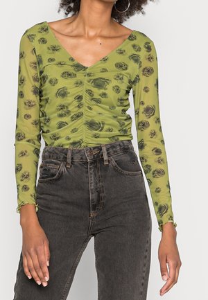 Vrouw draagt een groene doorschijnende top met lange mouwen en zwarte bloemenprint met geplooide voorkant, gecombineerd met hoge taille vervaagde zwarte jeans.
