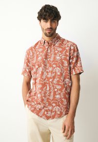 Chemise à manches courtes avec boutons, de couleur orange rouille avec un motif floral blanc, dotée d'un col et d'une poche poitrine, fabriquée en tissu léger.