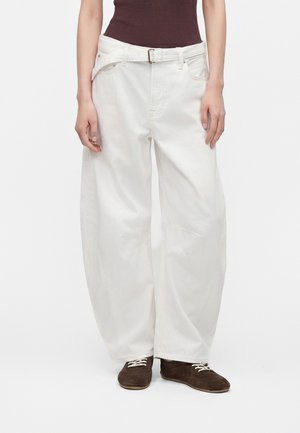 Persoon draagt losse witte jeans met een riem, een bruine geribde tanktop en donkerbruine veterschoenen, staand tegen een effen achtergrond.
